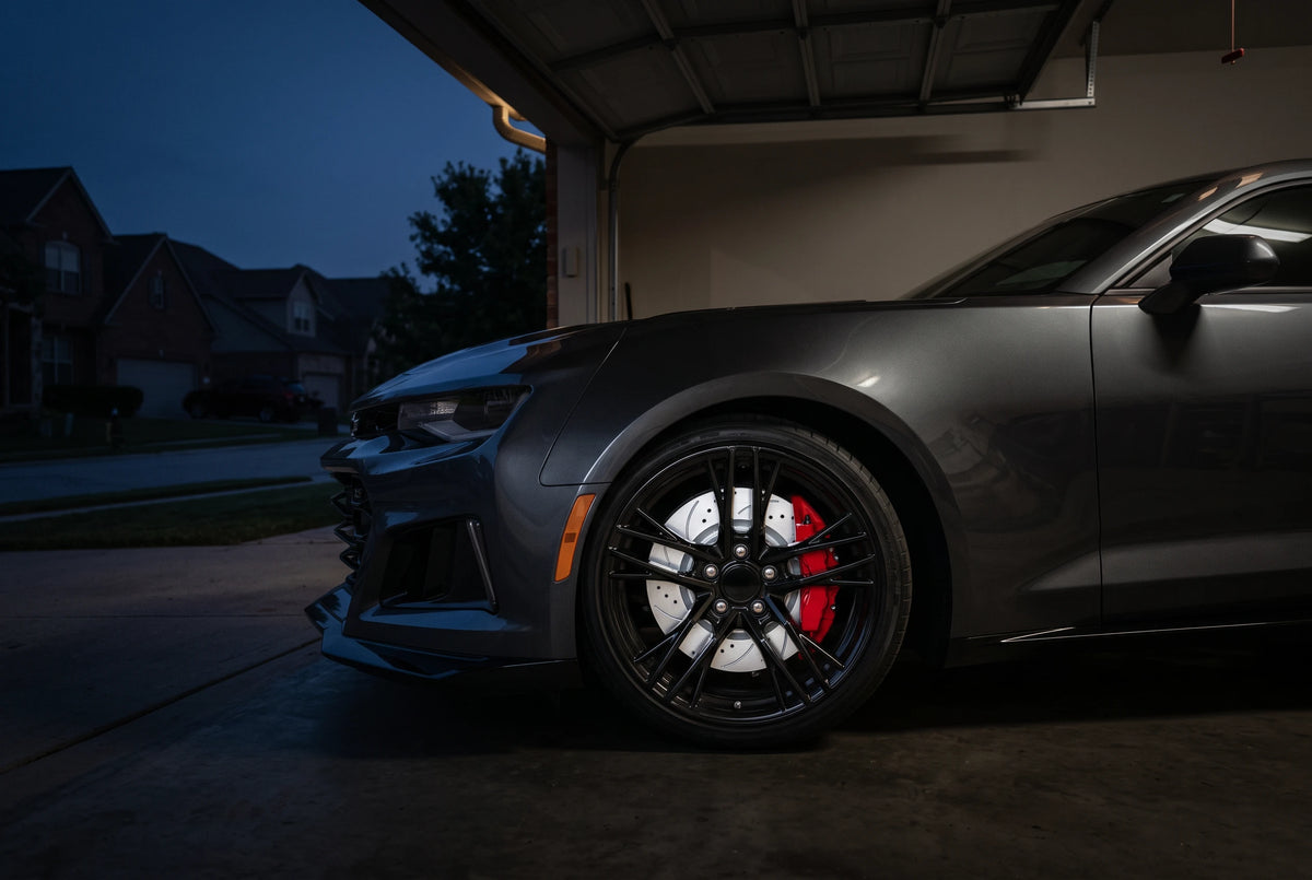 SP Performance double drilled and slotted brake rotor in silver zinc with a red caliper, on a dark gray car parked outside a home garage at dusk