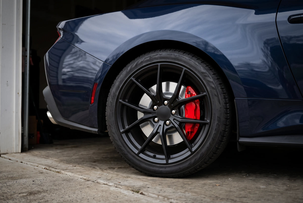 SP Performance plain brake rotor in silver zinc with a red caliper, behind the rear wheel of a blue car in a home garage