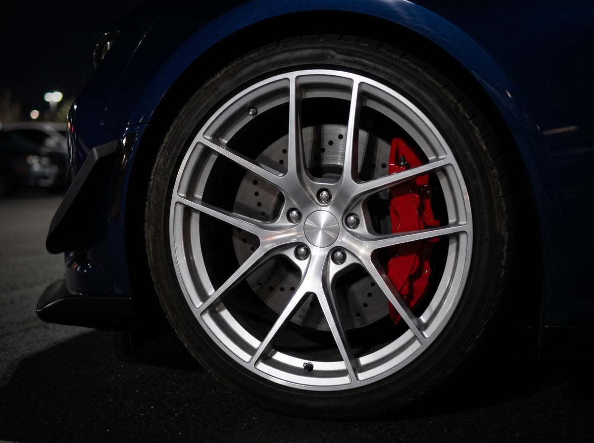 SP Performance cross drilled brake rotor in gray zinc with a red caliper, seen through the wheel of a blue Chevrolet Camaro ZL1 at a night car meet